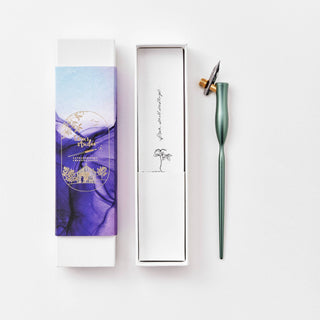The Flourish Calligraphy Pen, a green oblique pen with nib, rests by its open box featuring a blue and purple illustrated sleeve and an inner lid with a palm tree drawing and cursive text—ideal for beginner calligraphy.