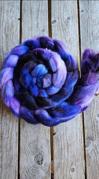 The Koigu Merino Roving Braid, hand-dyed in shades of purple, blue, and black, features a coiled design and is beautifully displayed in a spiral on a rustic wooden surface.