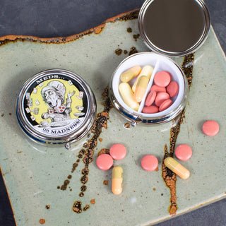 The Mad Hatter Stitch Marker / Pill Box with a cartoon “MEDS... OR MADNESS” is open to reveal red tablets and yellow capsules, plus scattered pills on a ceramic tray. Case features a mirrored lid—great for knitting accessories or stitch markers.