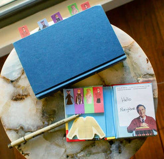 A blue book sits on a round stone table next to an open notebook with colorful page markers, a card with Hello Neighbor and Mister Rogers, and message-friendly Mister Rogers Sticky Notes shaped like sweaters, plus a pen.