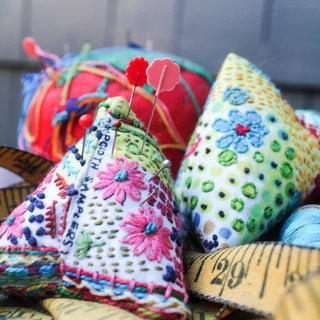 Colorful hand-embroidered pin cushions, some with pins, are shown with a measuring tape and thread—featuring vibrant floral designs to inspire your next creation with the Hand-Embroidered Pin Cushion Project Kit.