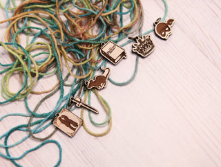 The Princess Bride Knitting Stitch Markers—five metal accessories shaped like a painting palette, cat, book, fire, and a person with a sword—are attached to multicolored yarn tangled on a white wooden surface.