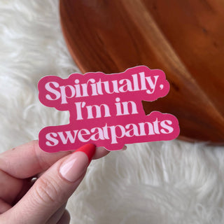 A hand with red and nude manicured nails holds the "Spiritually, I'm in sweatpants" sticker. The pink vinyl sticker features white lettering, with a fuzzy white surface and wooden object visible in the background.