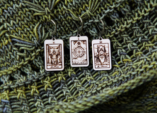 The 🔮 Tarot Card Stitch Markers – Justice, Wheel of Fortune & The Chariot 🔮 rest on a green and blue knitted background, perfect for pairing with crochet accessories or as unique stitch markers for your knitting projects.