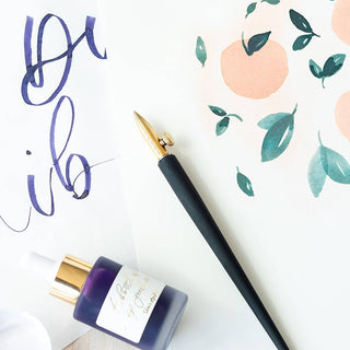 The One-Dip-Nib calligraphy pen with adjustable nib, a gold-capped dropper bottle, and two art sheets—watercolor peaches/leaves and purple calligraphy—are displayed on white, highlighting elegant line variation.