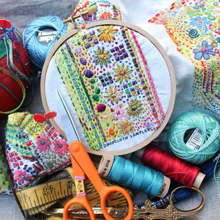 The Hand-Embroidered Pin Cushion Project Kit features a colorful, detailed embroidered hoop with floral patterns, surrounded by scissors, threads, a tape measure, and sewing tools on a work surface.