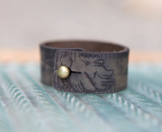 The Mushroom Leather Ruler Cuff, featuring brown leather with engraved mushroom designs and numbers, is fastened by a round brass stud and displayed on a textured blue and white surface.