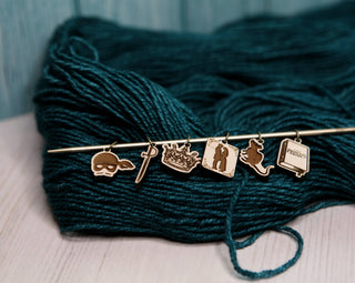 Resting on teal yarn atop a wooden surface, the Princess Bride Knitting Stitch Markers set includes six story-themed charms—a mask, sword, crown, dancing couple, mermaid, and open book—adorning a smooth maple wood knitting needle. These unique stitch markers add whimsical charm to your knitting accessories.