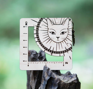 The Witchy Cat Gauge Swatch Ruler is a square maple wood keychain with a cat face, sunburst design, and engraved knitting gauge marks in cm and inches, shown balanced on wood against a blurred green background.