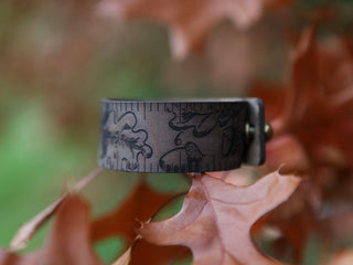 The Oak Leaves & Acorn Leather Ruler Cuff, made from brown reclaimed leather with engraved oak leaves and acorns, rests on reddish-brown oak leaves against a softly blurred green and brown background.