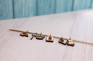 Six small, gold-colored Princess Bride Knitting Stitch Markers—shaped like a helmet, sword, crown, book, cat, and a second book—hang from a thin rod on a light maple wood surface with a blue wooden background. These whimsical charms are perfect knitting accessories or stitch markers for your next project.