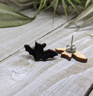 Two Bat Earrings rest on a wooden surface. One shows the black bat front, the other reveals the wooden back and metal post. Perfect handmade jewelry for Halloween, these accessories stand out against a backdrop of blurred green leaves.