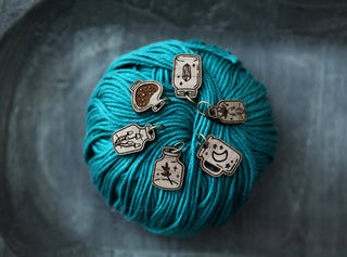 The Apothecary Bottle Stitch Marker Set features six walnut wood markers shaped like jars and mugs, each with unique engraving, paired with a ball of teal yarn—ideal knitting accessories for your next project.