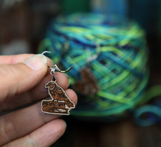 A hand displays the Conspiracy of Ravens Knitting Progress Keeper, featuring a brown bird design against a blurred backdrop of blue and green yarn—ideal inspiration for maple wood crochet accessories.