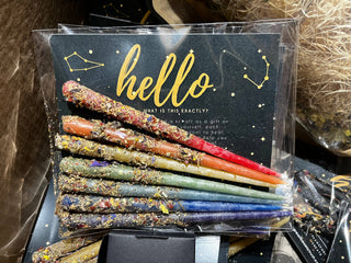 Kaleidoscope Beeswax Candles arranged in rainbow order on a black card with gold "hello" text, featuring star and constellation designs. The colorful candles are covered with dried herbs and have a vibrant, glittery appearance.