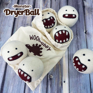 Six white Wool Felted Monster Dryer Balls, each with cute cartoon monster faces, are displayed on a wooden surface beside a WOOLBUDDY cloth bag with a dinosaur design—an eco-friendly, natural fabric softener for your laundry.