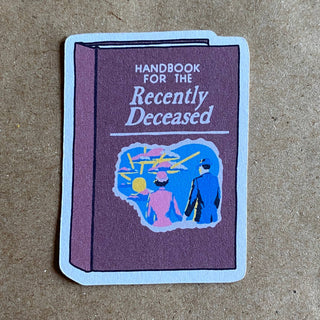 The Handbook for the Recently Deceased sticker shows a purple book cover with a vintage-dressed couple walking toward a bright sky. Made from eco-friendly paper, it stands out on a brown textured background.