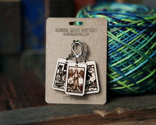 The 🔮 Tarot Card Stitch Markers – The Fool, Death & Temperance 🔮, ideal as crochet or knitting progress keepers, are shown on a Sunrise Grove Knittery backing with blue and green yarn on a wooden surface.