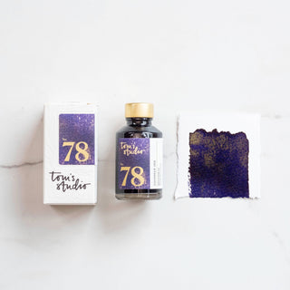 A bottle of Toms Studio Shimmer Ink - Broadway, its packaging box, and a swatch of dark purple fountain pen ink with gold shimmer are displayed on a white surface.