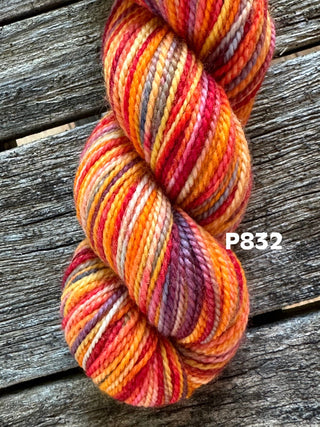 A skein from the KPM and KPPPM Variegated Color Collection in vibrant orange, yellow, red, and purple rests on weathered wood. "P832" appears on the right—ideal for your next hand knitting project.