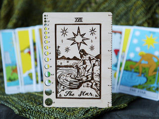 A Maple Wood Tarot Card Knitting Needle Gauge & 5 Ruler stands upright, engraved with The Star tarot design. In the background, colorful tarot cards are blurred on a green textured surface.