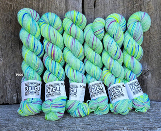 Six KPM and KPPPM Variegated Color Collection skeins in color P096, featuring light green with blue, purple, yellow, and pink speckles, are arranged before a rustic wooden background—ideal for hand knitting projects.