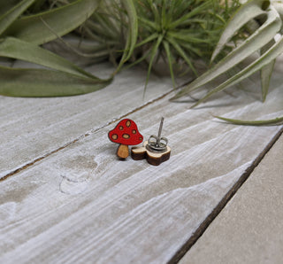 A charming, handmade-in-USA Mushroom Earring—small, red, with white spots—rests on a light wooden surface beside its metal backing and lush green leaves.