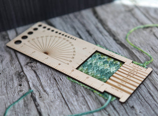 The Butterfly "Almost Everything" Ruler, a wooden knitting and crochet tool with measurement markings, sits on gray wood over green and blue knitted fabric and yarn—ideal for easily checking stitch size and yarn weight.