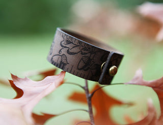 The Oak Leaves & Acorn Leather Ruler Cuff, made from brown reclaimed leather and engraved with oak leaves and acorns, rests on orange autumn leaves against a blurred green background.