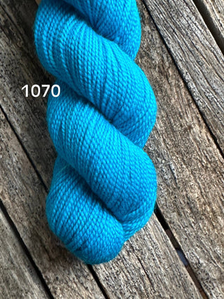 A skein of Koigu KPM & KPPPM Fingering Weight Yarn in bright turquoise blue (color 1070) is displayed on a rustic wooden surface featuring weathered, textured planks.