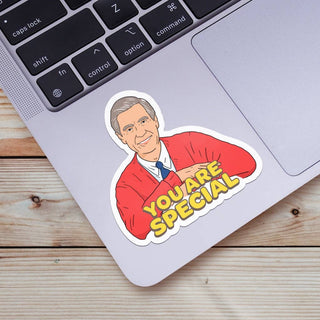 The "You Are Special | Mr. Rogers Sticker"—a high-quality vinyl of a smiling man in a red cardigan with “YOU ARE SPECIAL”—is displayed on a silver laptop by the keyboard, promoting mental health awareness on a wooden desk.