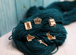 Six Princess Bride Knitting Stitch Markers—including a couple silhouette, a crown, a “Princess” book, a mask, a sword, and a mermaid—sit atop teal yarn and are beautifully arranged on a light maple wood background.