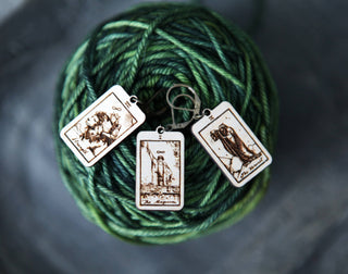 The 🔮 Tarot Card Stitch Markers – The Hermit, The Magician & Strength 🔮 feature three intricate rectangular charms, perfect as stitch markers or progress keepers. Shown on green and blue variegated yarn with a softly blurred gray background.