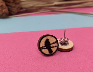 Crow Earrings: Handmade wooden earrings engraved with a crow silhouette, displayed on a pink and blue surface, with dried plants softly blurred in the background.