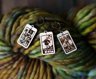 Three rectangular 🔮 Tarot Card Stitch Markers – The Fool, Death & Temperance 🔮 rest on chunky multicolored yarn, making unique stitch markers or knitting progress keepers for your creative projects.