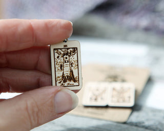A hand holds a maple wood "The Tower" pendant from the Tarot Card Stitch Markers–The Tower, The Devil & Judgement set, designed as a unique knitting progress keeper. Two more tarot-themed markers are blurred in the background.