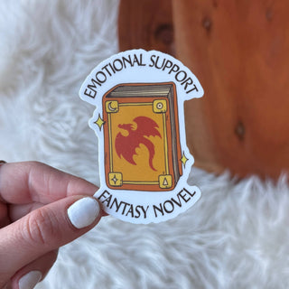 Sticker with 'Emotional Support Fantasy Novel' design held by a hand on a soft white background.