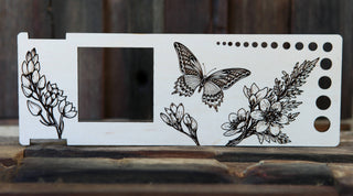 The Butterfly "Almost Everything" Ruler is a rectangular wooden knitting gauge with a cut-out square, circular holes, and detailed black line drawings of flowers and a butterfly on a rustic wood background.