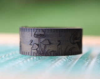 A close-up of the Mushroom Leather Ruler Cuff, featuring engraved ruler markings, numbers, and detailed mushroom illustrations, set against a blurred green background.