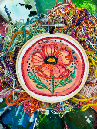 A pink flower with green leaves, black and yellow accents from the Cosmic Poppy Embroidery Kit is embroidered on a nest of colorful threads with splashes of paint in the background.