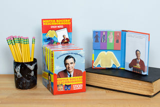 A set of Mister Rogers Sticky Notes, shaped like his iconic sweater, is displayed on a table beside a mug of pencils and a large closed book, with one pad open to show the notes and a photo of Mister Rogers.