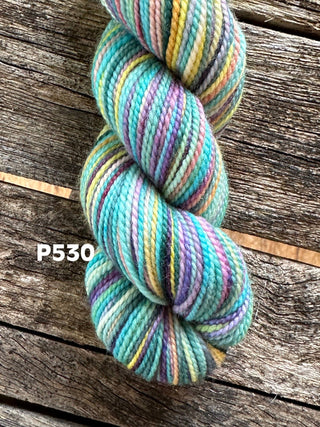 A skein from the KPM and KPPPM Variegated Color Collection in teal, purple, yellow, and green rests on rustic wood. The soft merino wool yarn features the label P530 in white text—ideal for hand knitting projects.