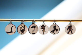 The 👽 I Believe Stitch Marker Set 👽 features six circular maple wood stitch markers etched with Nessie, Mothman, UFO abduction, fairy, unicorn, and Bigfoot—unique knitting accessories on a blurred blue and white background.