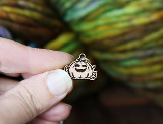 A hand holds a 🎃 Pumpkin Jack O’Lantern Stitch Marker, a small wooden pin shaped like a smiling pumpkin with carved eyes and mouth, set against a blurred, colorful background.