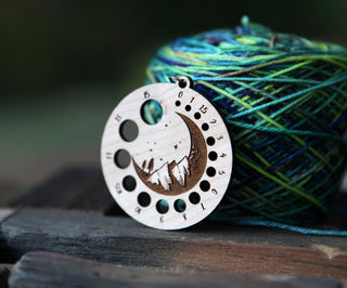 The Crescent Moon Knitting Needle Gauge, crafted from cherry wood and engraved with a moon, mountains, and trees, is shown in front of blue and green multicolored yarn on a wooden surface.
