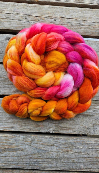 The Koigu Merino Roving Braid, made of thick merino wool in vibrant orange, yellow, pink, and red shades, is coiled in a spiral on a weathered wooden surface.