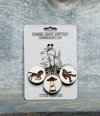 The 👽 I Believe Stitch Marker Set 👽 features three maple wood markers on a card, engraved with a wolf, Bigfoot, and a UFO. Card reads Sunrise Grove Knittery with website & "Stitch Markers." Unique knitting accessories for your project!.