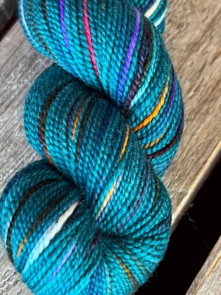 A close-up of a KPM and KPPPM Variegated Color Collection skein in teal with streaks of purple, orange, white, and black rests on weathered wood—perfect for your next hand knitting project.