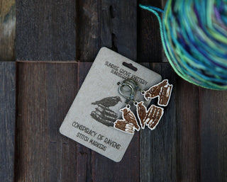 Conspiracy of Ravens Knitting Progress Keepers are attached to a labeled card on a rustic wood surface, perfect as maple wood stitch markers or crochet accessories. A colorful ball of yarn sits nearby.