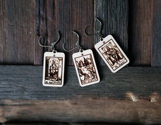 Three maple wood stitch markers, engraved with The High Priestess, The Empress & The Emperor tarot cards, hang against rustic wood. These Tarot Card Stitch Markers are perfect progress keepers for knitting and crochet enthusiasts.
